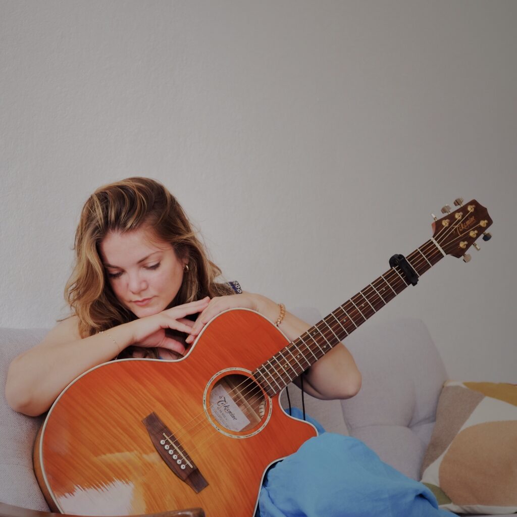 Frau sitzt auf einem Sofa mit einer orangefarbenen Gitarre auf dem Schoß. Der Hintergrund ist grau