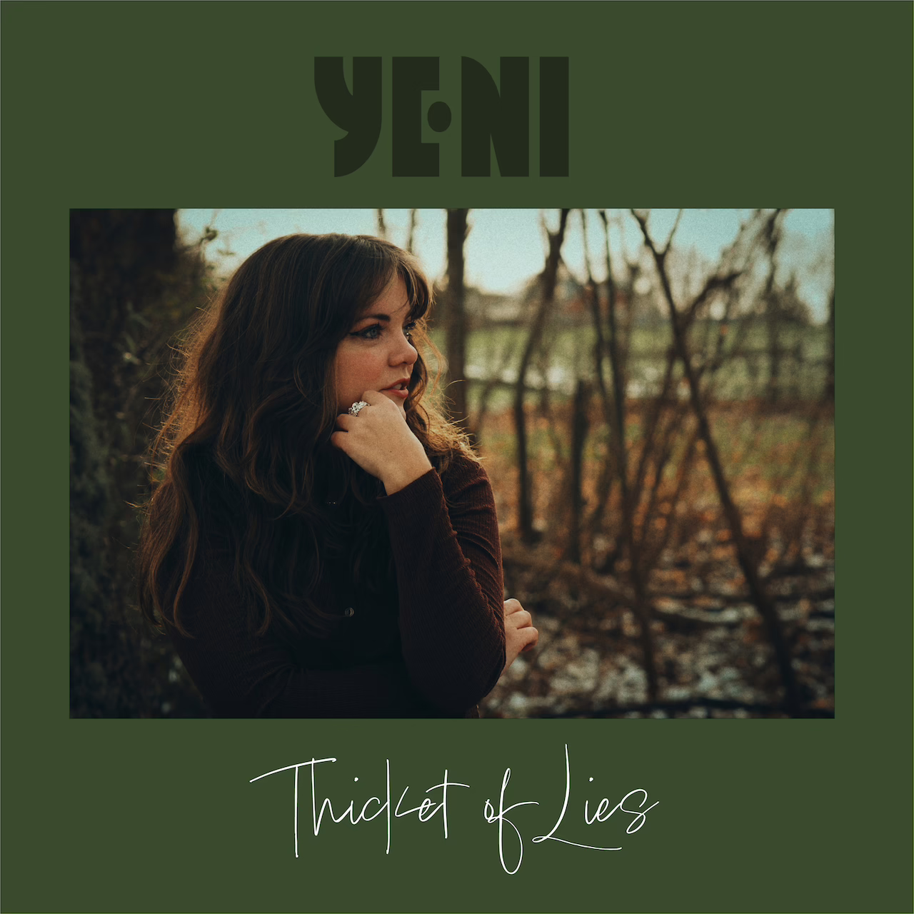 Single cover artwork for Thicket of lies with a photo of YENI dressed in a dark brown shirt, looking to the side with the hand under her chin. the surrounding area is green and above the photo is the YENI logo in a darker shade of green and below is the text Thicket of Lies in a cursive white font