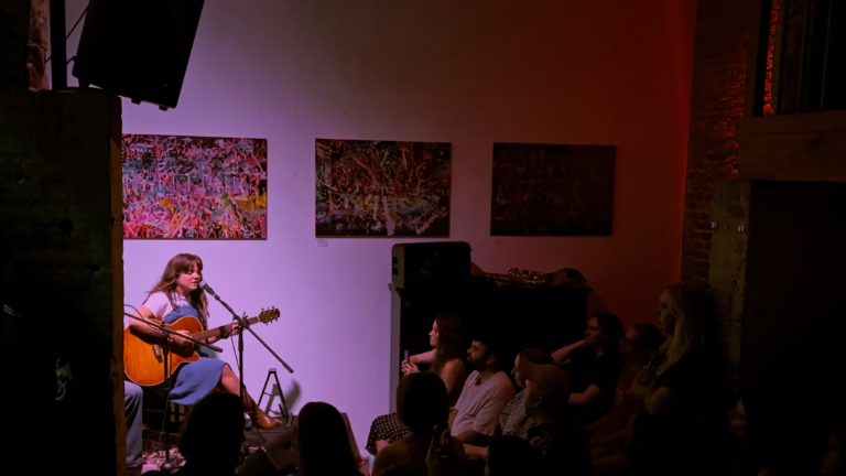 Wide shot of YENI onstage, she is sitting on a chair, playing guitar and singing with her eyes closed. The audience is listening attentively.