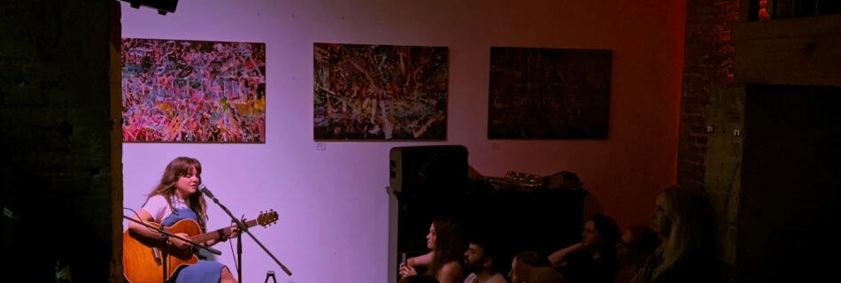 Wide shot of YENI onstage, she is sitting on a chair, playing guitar and singing with her eyes closed. The audience is listening attentively.